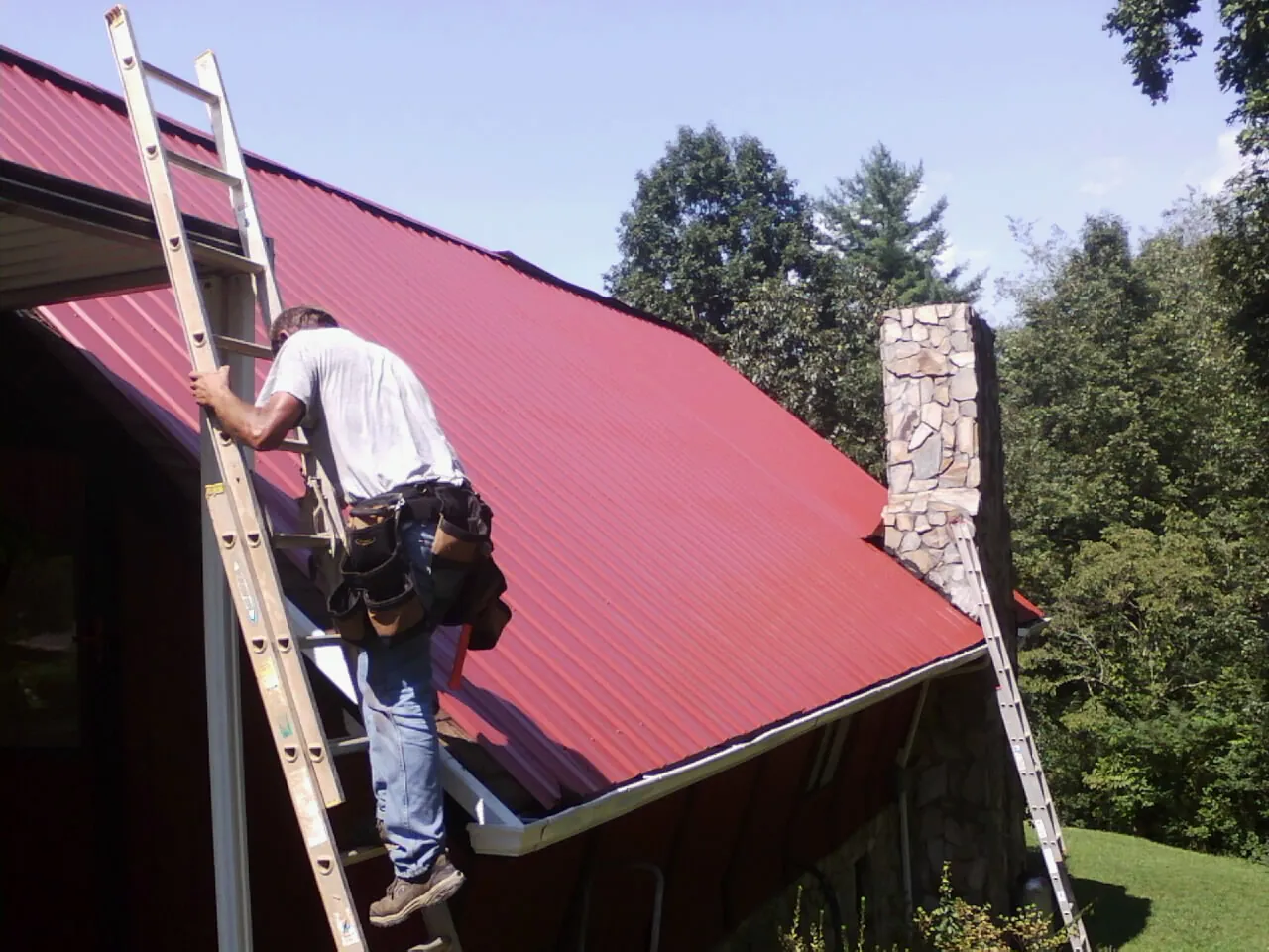 Roofer on a ladder beside a red metal roof and stone chimney — Soffit & Fascia Repair by Roofing Brothers in Davilla